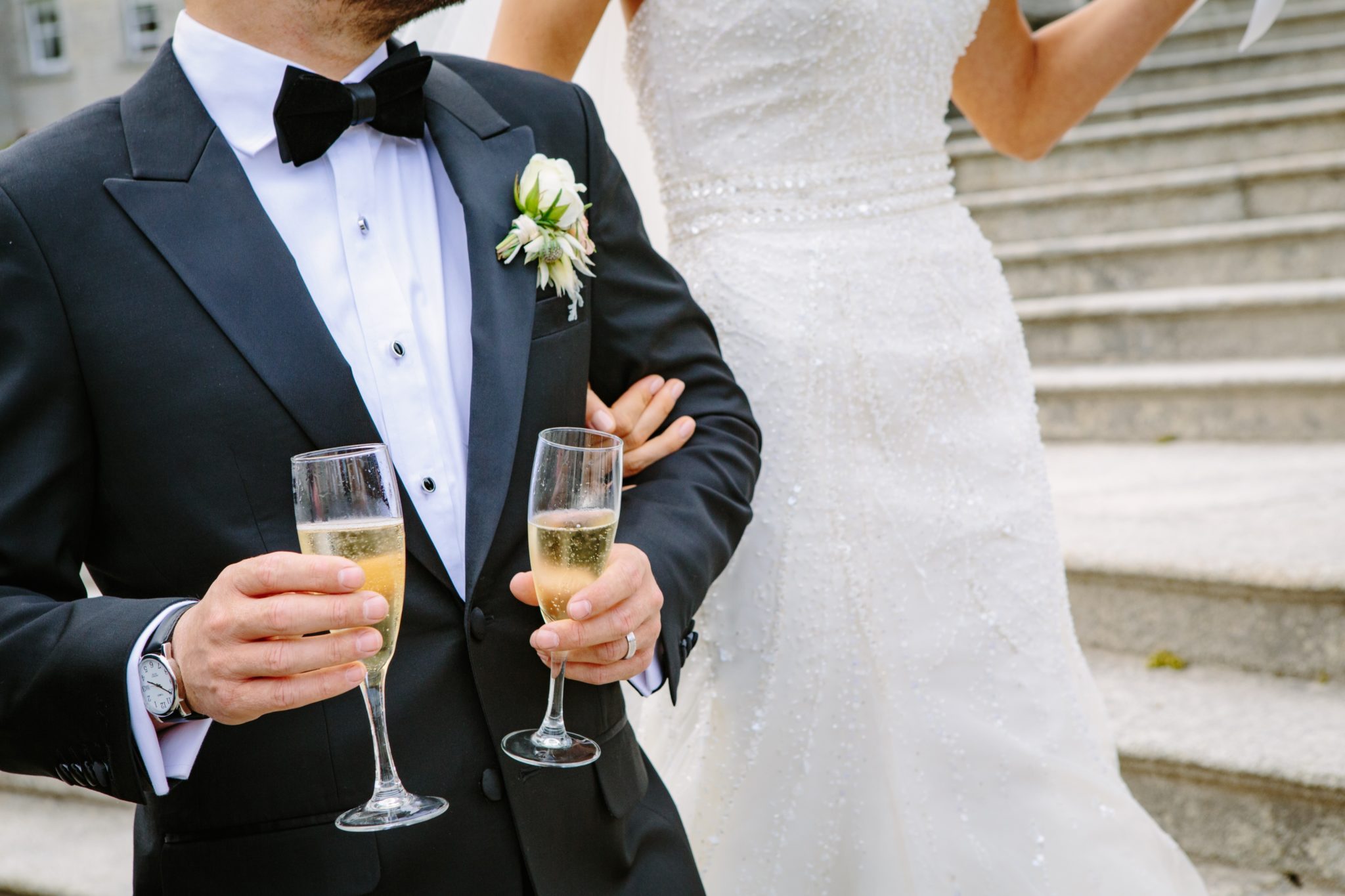 Groom wearing tuxedo holding champagne glasses with bride on his arm