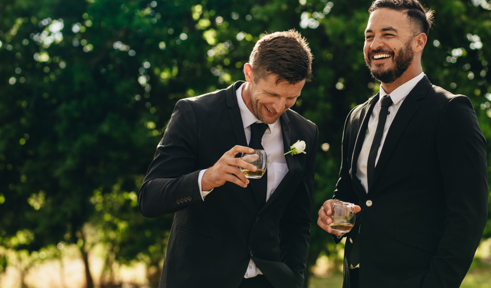 Groom and brother in black suits laugh while sipping on a cocktail at outdoor wedding
