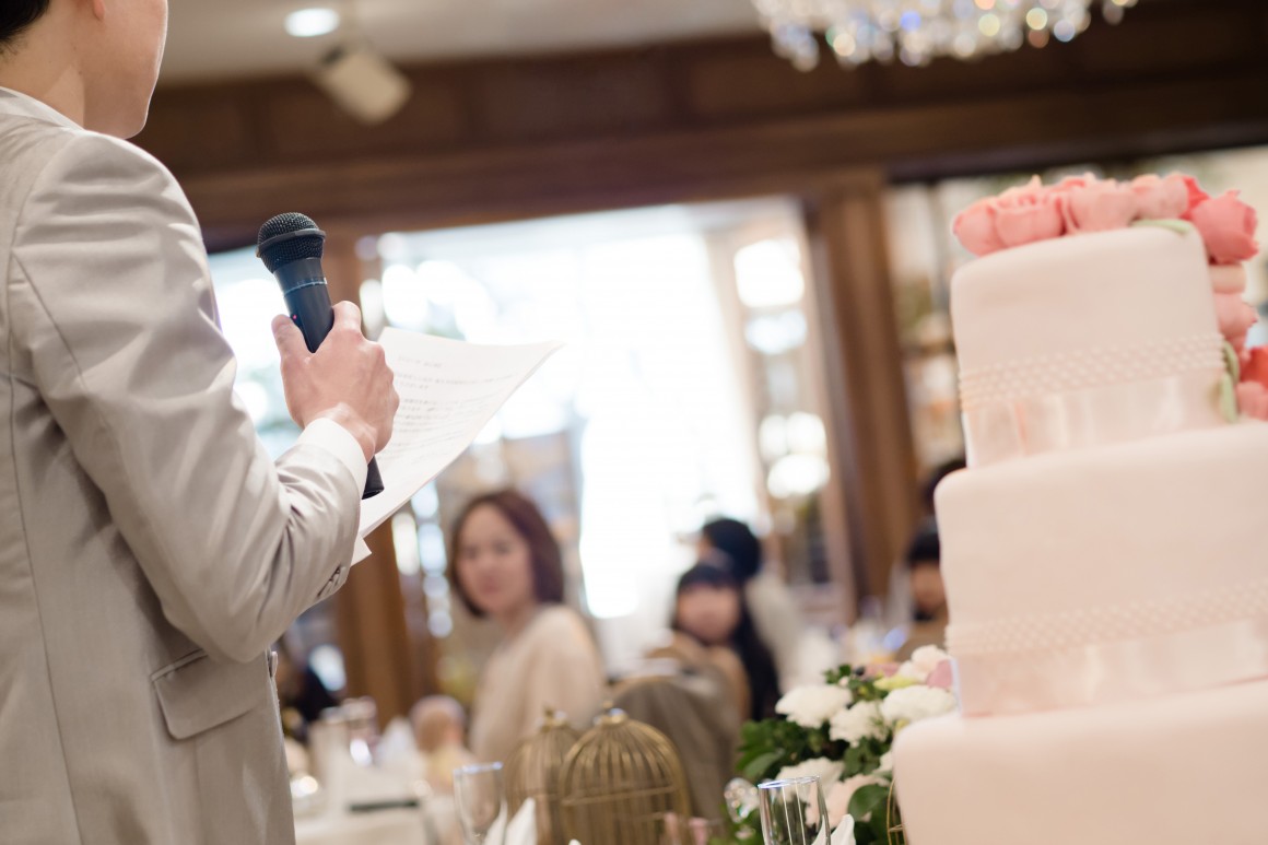 Groom in tan suit standing next to ivory wedding cake delivering speech.